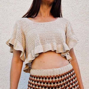 May include: A cream-coloured, crocheted crop top with ruffled sleeves and a matching skirt. The top has a round neckline and a ruffled hem. The skirt features a geometric pattern in shades of brown and cream. The outfit is handmade.
