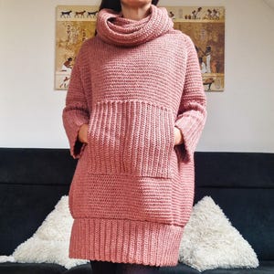 Può includere: Un maglione lungo rosa lavorato a uncinetto con collo alto e una grande tasca sul davanti.