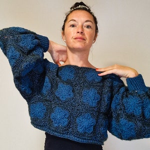 Peut inclure: Un pull en crochet bleu et vert avec un motif floral texturé. Le pull est court et à manches longues.