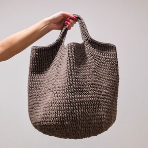Puede incluir: Un bolso tote grande de color marrón tejido a crochet con un solo asa. El bolso está hecho de un hilo grueso y texturizado y tiene un interior espacioso.