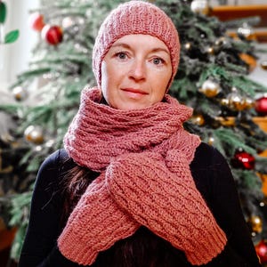 May include: A pink knitted hat, scarf, and mittens set. The hat has a ribbed texture and the scarf and mittens have a cable knit pattern.