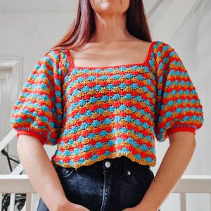 Puede incluir: Un top de crochet con escote cuadrado y mangas abullonadas cortas. El top está hecho con un patrón de rayas coloridas en tonos rojos, naranjas, azules y marrones.