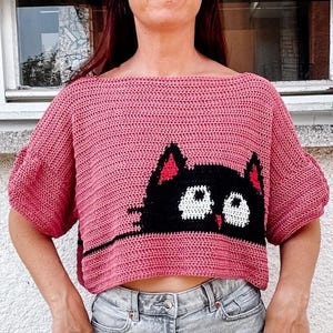 May include: A pink crocheted crop top with a black cat face design. The cat has white eyes and a red nose.