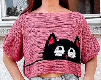 May include: A pink crocheted crop top with a black cat face design. The cat has white eyes and a red nose.
