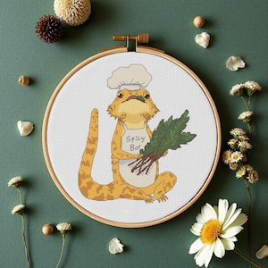 Può includere: Un ricamo a punto croce in un telaio di legno rotondo raffigurante un drago barbuto giallo che indossa un cappello da chef e un grembiule bianchi. Il grembiule riporta la scritta Spicy Boi. La lucertola tiene in mano verdure a foglia verde.