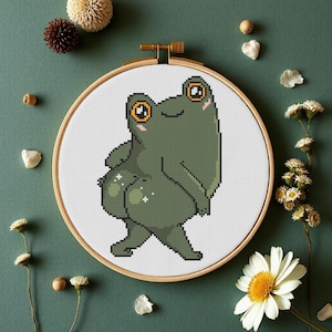 May include: A green frog with big eyes and a smile is embroidered on a white canvas in a wooden embroidery hoop. The frog is standing on its hind legs with its front legs crossed.