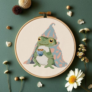 Puede incluir: Una rana verde con un sombrero rosa y azul y una manta rosa y azul, sosteniendo una pequeña taza de café. La rana está sentada sobre un fondo blanco dentro de un aro de bordado de madera.