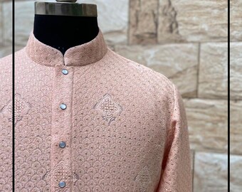 Op de afbeelding: Een licht perzikgekleurd, geborduurd kurta-shirt met een mandarijnkraag en knoopsluiting. Het shirt heeft ingewikkelde patronen en decoratieve stiksels. Het kledingstuk wordt op een mannequin getoond.