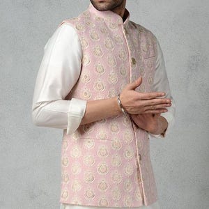 May include: A light pink embroidered vest with a mandarin collar and gold buttons. The vest is worn over a white long-sleeved shirt. The vest features a repeating floral pattern.