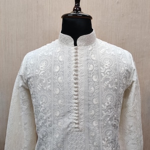 May include: Cream-coloured long-sleeved tunic with detailed floral embroidery. The tunic has a mandarin collar and a button-down placket. The fabric appears lightweight, with a detailed pattern. The garment is displayed on a mannequin.