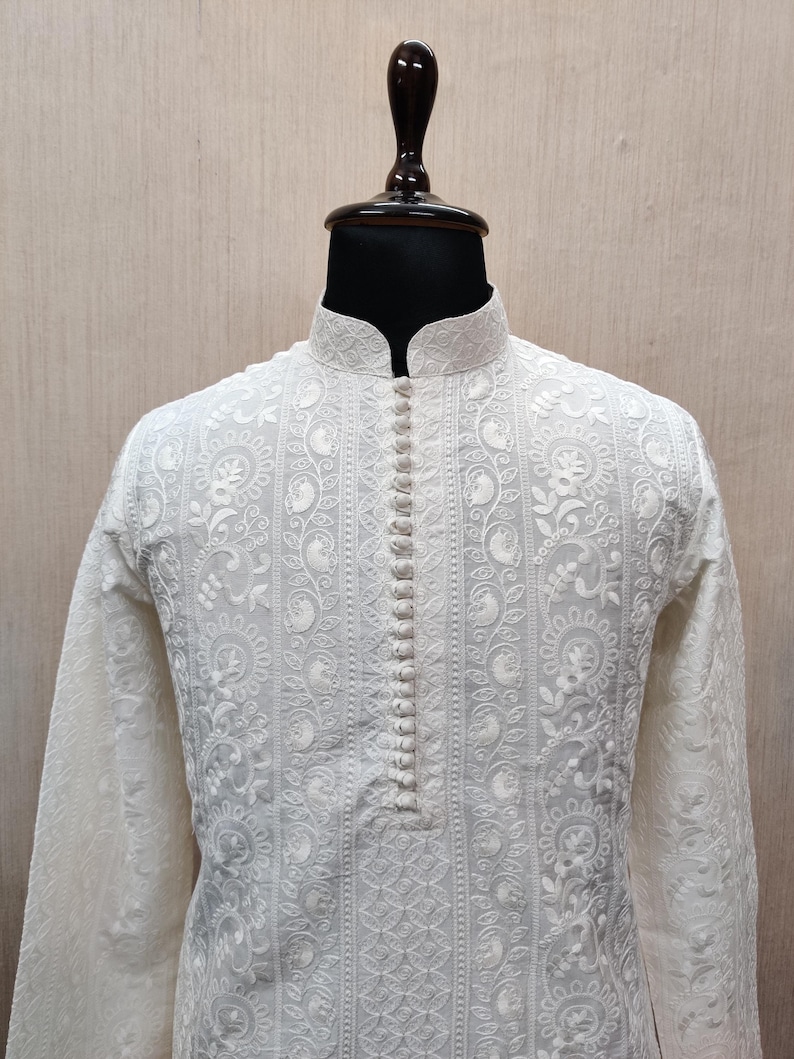 May include: Cream-coloured long-sleeved tunic with detailed floral embroidery. The tunic has a mandarin collar and a button-down placket. The fabric appears lightweight, with a detailed pattern. The garment is displayed on a mannequin.