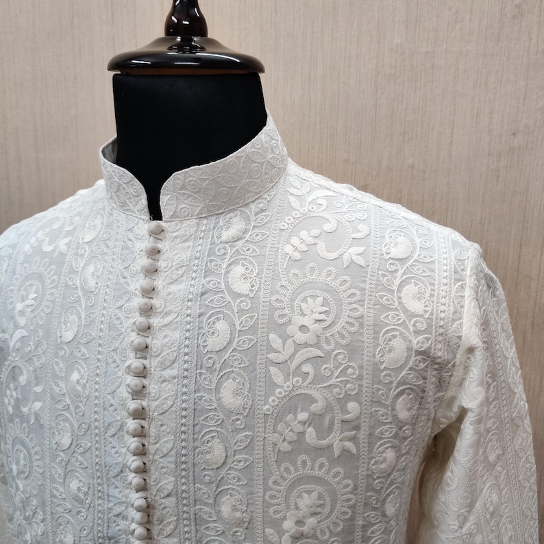 May include: White embroidered shirt with a mandarin collar and button-down front. The shirt features intricate floral and paisley patterns. The long-sleeved shirt is displayed on a mannequin.