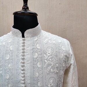 May include: A white, long-sleeved shirt with detailed floral embroidery and a mandarin collar. The shirt has a button-down front and ornate patterns, displayed on a mannequin.