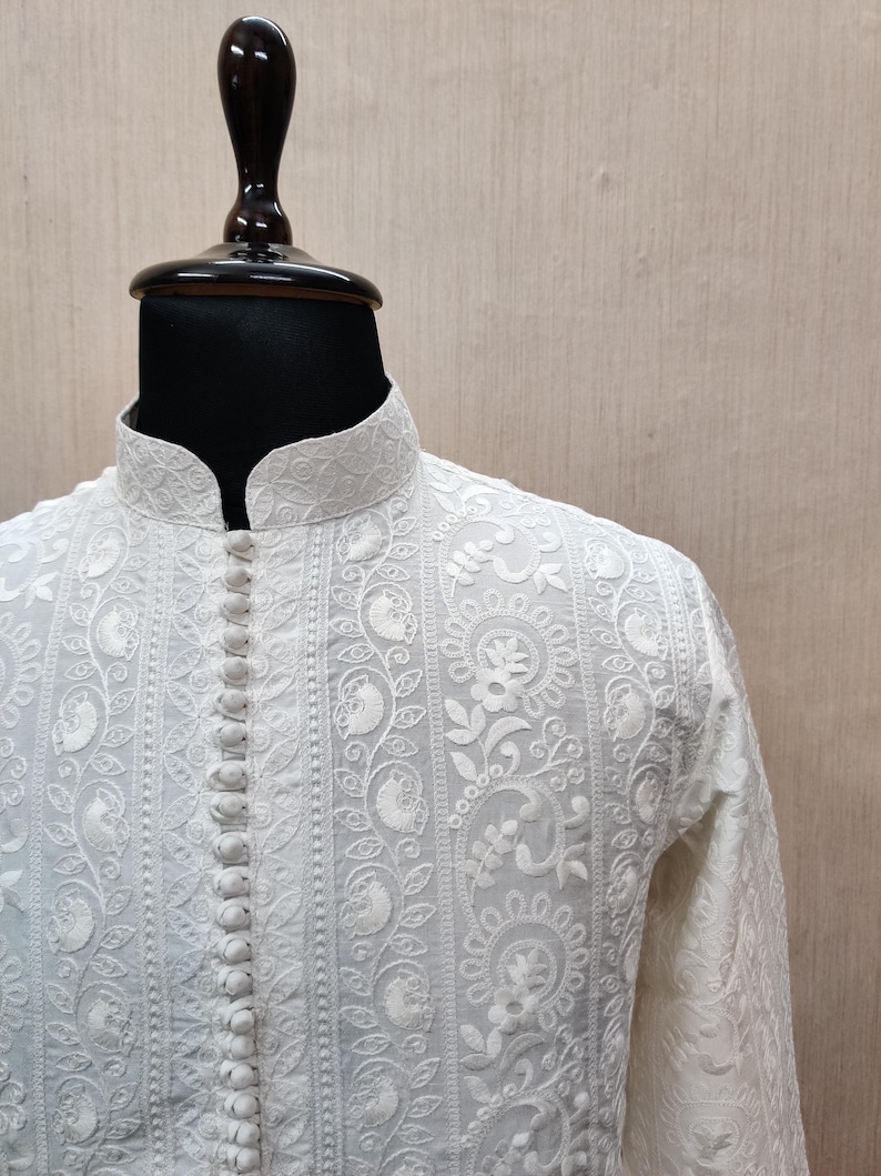 May include: A white, long-sleeved shirt with detailed floral embroidery and a mandarin collar. The shirt has a button-down front and ornate patterns, displayed on a mannequin.