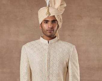 May include: A man wearing a cream-colored turban with a decorative feather and gold embellishment. He is also wearing a cream-colored jacket with a detailed, repeating pattern. The background is a solid, neutral color.