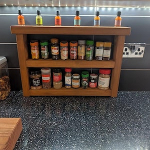 May include: A three-tiered wooden spice rack, holding numerous spice jars and small bottles. The rack is made of light brown wood and is placed on a black countertop. The spices are in clear glass jars with colourful labels.