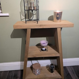 May include: A light oak side table with two shelves. A black metal lamp with a glass dome sits on the top shelf, alongside a lit candle in a gold container. The lower shelves hold a pink wax warmer and a glass jar with contents.