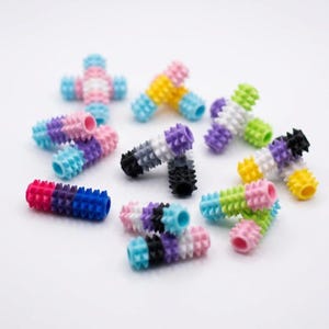 May include: A collection of small, cylindrical silicone beads featuring a textured, spiky surface. Each bead is composed of multiple colored segments arranged in various patterns, including pink, blue, white, yellow, purple, black, and green.