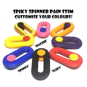 Peut inclure: Plusieurs jouets fidget ovales et colorés avec des centres circulaires texturés. Les jouets sont de différentes couleurs, dont le jaune, le rose, l'orange, le violet, le bleu, le noir et le rouge. Le texte sur l'image indique "SPIKY SPINNER PAIN STIM CUSTOMISE YOUR COLOURS!"