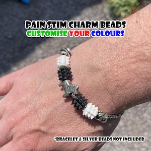 Pode incluir: Uma pulseira com contas pretas, brancas e prateadas. A pulseira tem um design de corrente com várias formas de contas, incluindo contas pontiagudas e em forma de estrela. O texto na imagem diz "PAIN STIM CHARM BEADS CUSTOMISE YOUR COLOURS".