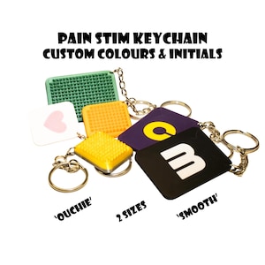 May include: A selection of keychains in diverse colours and textures, including green, yellow, purple, white, and black. Each keychain features a metal ring and chain. Text on the keychains reads "PAIN STIM KEYCHAIN CUSTOM COLOURS & INITIALS", "OUCHIE", "2 SIZES", and "SMOOTH".