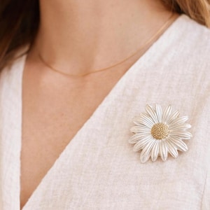 May include: A silver-toned daisy brooch with a gold-colored center is pinned to a white textured fabric. The brooch is a detailed floral design, showcasing delicate petals. A gold necklace is also visible.
