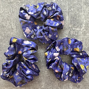 Op de afbeelding: Drie blauwe satijnen scrunchies met een Halloween-thema. De scrunchies bevatten geesten, pompoenen en andere Halloween-afbeeldingen.
