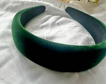 May include: A padded headband covered in emerald green velvet. The headband has a rounded shape and a dark green interior. The accessory is displayed on a white, textured surface, with a few flowers in the background.
