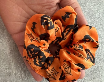 May include: Orange and black Halloween-themed scrunchie with a pattern of pumpkins, bats, stars, and spiderwebs.