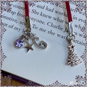 Puede incluir: Un marcapáginas rojo con abalorios plateados. Los abalorios incluyen una estrella, una letra G y un cristal morado. El marcapáginas está unido a un libro con el texto "She knew what anyone. Charlie Ja, es wa To me she was attain".