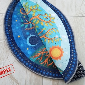 May include: A round rug with a blue and green tree of life design. The rug has a dark blue border with white dots. The rug is folded in half, showing the back side which is gray with a black non-slip backing.