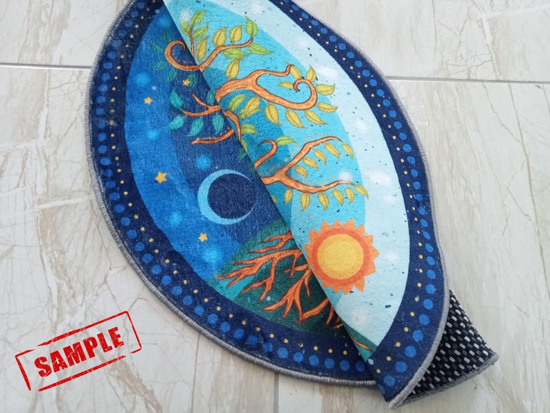 May include: A round rug with a blue and green tree of life design. The rug has a dark blue border with white dots. The rug is folded in half, showing the back side which is gray with a black non-slip backing.