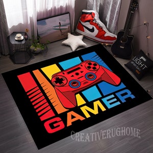 May include: A black and colorful gamer rug with a red and blue video game controller graphic and the word "GAMER" in rainbow colors. The rug has a black background with colorful stripes.