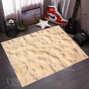 May include: A rectangular rug with a sand design, featuring beige and tan tones, creating a beach-like appearance. The rug has a textured surface with footprints, adding to the realistic look. The rug is placed on a dark wooden floor.