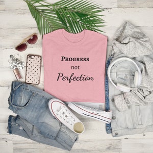 Może przedstawiać: Różowa koszulka z napisem "Progress not Perfection" czarnymi literami. Koszulka leży na białym drewnianym tle z dżinsami, dżinsową kurtką, parą białych butów sportowych, okularami przeciwsłonecznymi i słuchawkami.