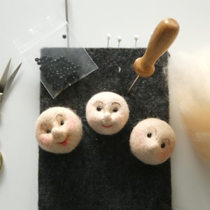 May include: A craft scene with three felted doll faces with painted features, a bag of black beads, scissors, a needle tool, and a pile of wool. The faces are on a dark gray felt square.
