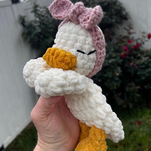 K&ouml;nnte beinhalten: Geh&auml;kelte Enten-Pl&uuml;schfigur mit wei&szlig;em K&ouml;rper, gelben F&uuml;&szlig;en und Schnabel sowie einer rosa Schleife. Die Stoffente hat geschlossene Augen und besteht aus strukturiertem Garn.