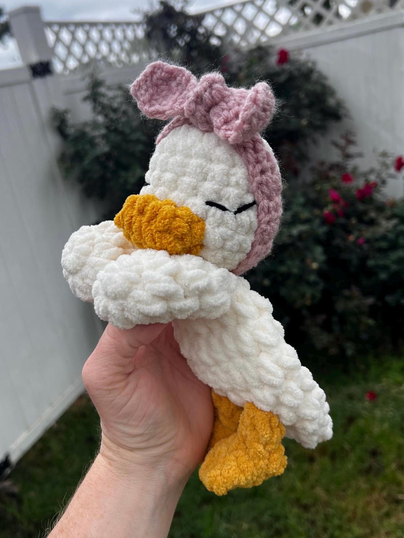 K&ouml;nnte beinhalten: Geh&auml;kelte Enten-Pl&uuml;schfigur mit wei&szlig;em K&ouml;rper, gelben F&uuml;&szlig;en und Schnabel sowie einer rosa Schleife. Die Stoffente hat geschlossene Augen und besteht aus strukturiertem Garn.