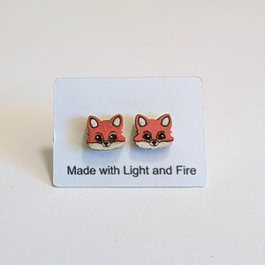 May include: A pair of wooden fox-shaped earrings. The earrings are orange with white and black details, and are displayed on a white card. The card has the text "Made with Light and Fire".