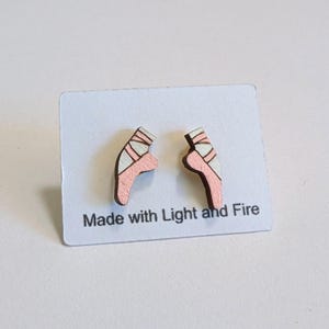 May include: Wooden ballet slipper earrings in pink and white, displayed on a white card. The earrings are shaped like ballet shoes with pink bodies and white accents. The card reads "Made with Light and Fire."