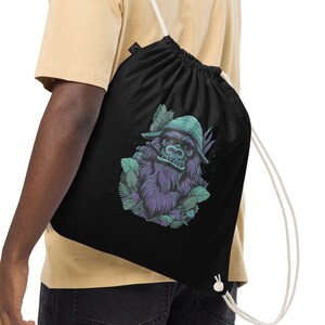 Op de afbeelding: Een zwarte trekkoordtas met een wit trekkoord. De tas heeft een afbeelding van een groene en paarse gorilla die een hoed draagt en omringd is door groene bladeren.