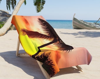 May include: A beach towel featuring a sunset and palm tree silhouette design. The towel is draped over a wooden beach chair on a sandy beach.