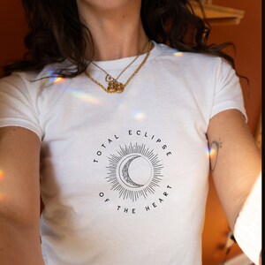 Könnte beinhalten: Weißes, bauchfreies T-Shirt mit einem schwarzen Motiv eines zunehmenden Mondes und einer Sonne mit dem Text "Total Eclipse of the Heart".