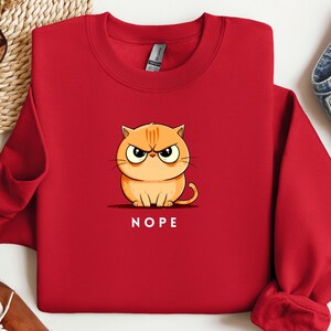 May include: Red sweatshirt with a cartoon orange cat with an angry expression and the word "NOPE" printed below it.