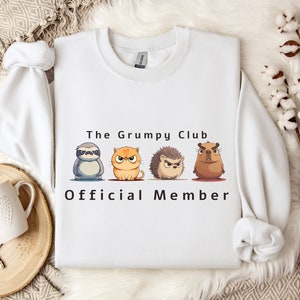 May include: White sweatshirt with the text "The Grumpy Club Official Member" and four cartoon animals: a sloth, a cat, a hedgehog, and a capybara.