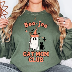 May include: Green sweatshirt with a ghost wearing a witch hat and holding a broom. The text "Boo-jee Cat Mom Club" is printed on the front of the sweatshirt.