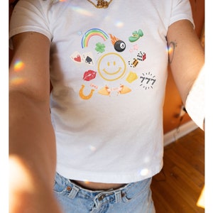 Könnte beinhalten: Weißes T-Shirt mit einem grafischen Druck eines Smiley-Gesichts, umgeben von verschiedenen Glücksbringer, darunter ein Regenbogen, ein vierblättriges Kleeblatt, ein Hufeisen, Würfel, eine Spielkarte, 777 und eine schwarze Kugel.