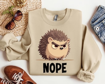 Könnte beinhalten: Beiges Sweatshirt mit einem Cartoon-Igel-Motiv. Der Igel hat einen wütenden Gesichtsausdruck und stachelige Stacheln. Das Wort "NOPE" ist in Schwarz darunter gedruckt. Sonnenbrille und eine gewebte Matte sind im Hintergrund.