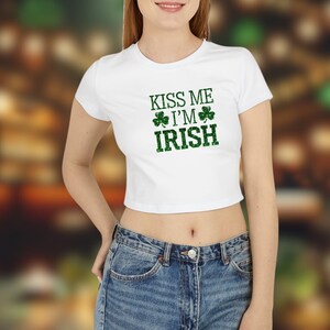 Könnte beinhalten: Weißes Crop-Top mit dem grünen Text "KISS ME I'M IRISH" und Kleeblatt-Grafiken. Das Top wird mit blauen Jeans getragen. Der Hintergrund ist verschwommen, was auf eine unscharfe Umgebung hindeutet.