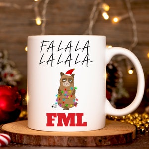 Puede incluir: Taza de cerámica blanca con un gato gruñón que lleva un gorro de Papá Noel y está enredado en luces de Navidad. El texto "Falala Lalala... FML" está impreso en la taza.